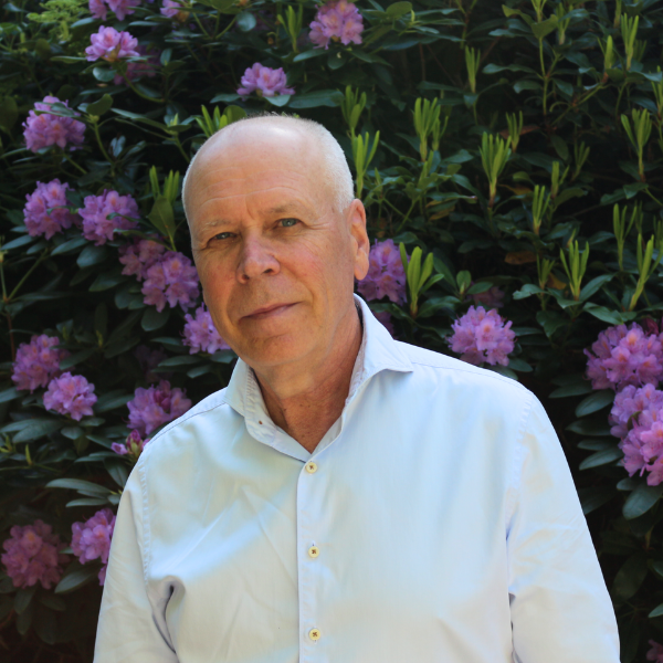 Äldre manlig terapeut i Alingsås, klädd i ljusblå skjorta, står framför blommande lila rhododendron i dagsljus – JiO Samtal och Psykoterapi